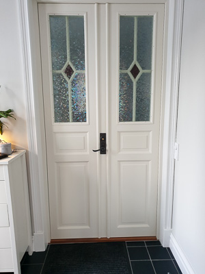 Vit pardörr med 2 höga glasrutor och spröjs i diamantmönster, med färgad glasdetalj som fångar ljuset i entrén.