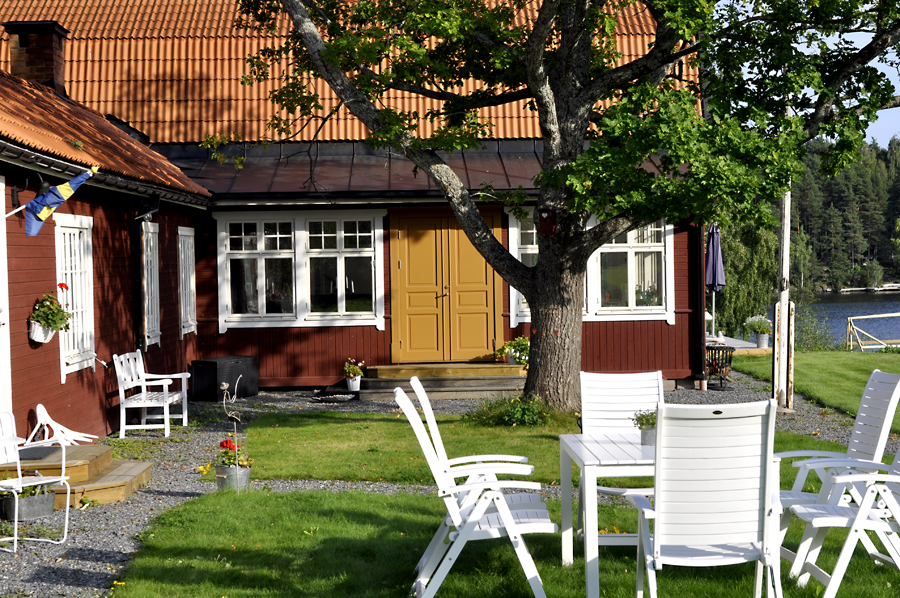 Rött trähus med gul pardörr och vitmålade spröjsade fönster, omgivet av gräsmatta, träd och vita trädgårdsmöbler nära vatten.