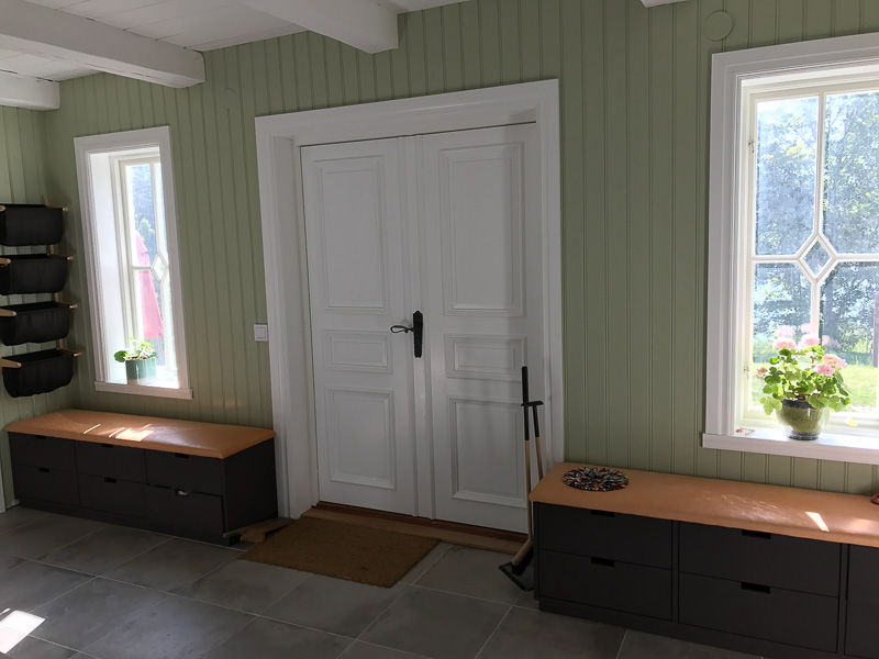 Interiör med vita dubbeldörrar och två smala fönster med romb/diamant-detalj i glaset, gröna väggpaneler och bänkar under fönstren.