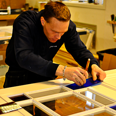En hantverkare lutar sig koncentrerat över ett fönster i verkstaden och skär till antikglas med hjälp av en glasskärare och en blå mall. Arbetet utförs med stor precision när glaset anpassas till spröjsindelningen i den målade träramen, som en del av traditionellt fönsterhantverk.