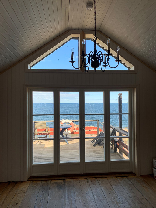 Spröjsade fönsterdörrar i ljus karm med stora glasytor och utsikt mot altan och vatten, med gavelöverljus ovanför.