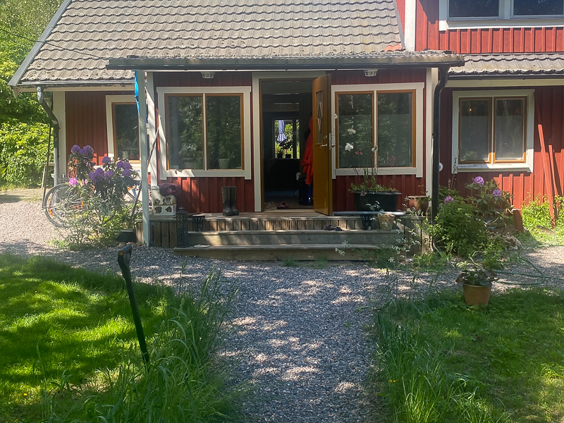 Fasad före ombyggnad med större fönsterpartier i vit karm som speglar grönska och dagsljus.