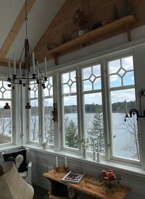 Hörnparti med vita fönster i vinkel, där de höga rutorna har dekorativa rombdetaljer upptill och ger panoramakänsla mot sjö och träd.
