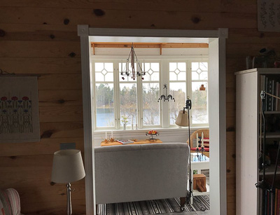 Vy genom en dörröppning mot en ljus fönstervägg med höga vita fönster och dekorativa rombdetaljer i överkant, där sjö och skog syns genom glasen.