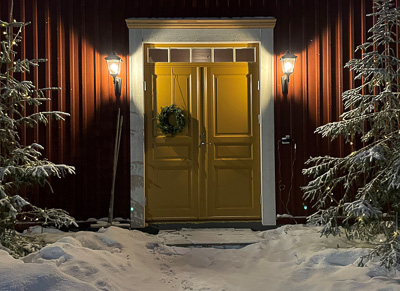 Gul pardörr i röd träfasad med rektangulärt överljusfönster, där glaset fångar ljuset från entrébelysningen i vintermörker.