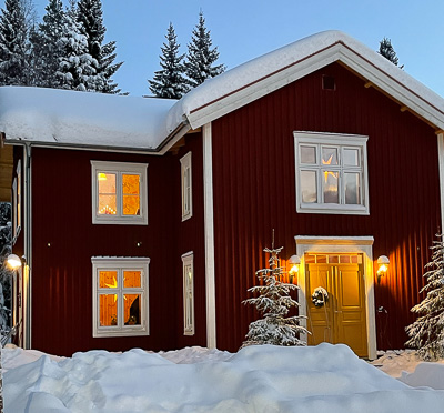 Rött hus i vinterkväll med vitmålade fönster som lyser varmt inifrån, samt gul pardörr med överljusfönster som släpper in ljus i entrén.
