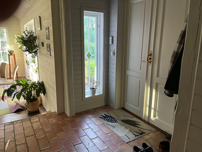Hall/entré med vit ytterdörr och ett smalt, högt sidofönster med dekorativ rombform i glaset, där dagsljuset faller in och grönskan utanför syns genom rutan.