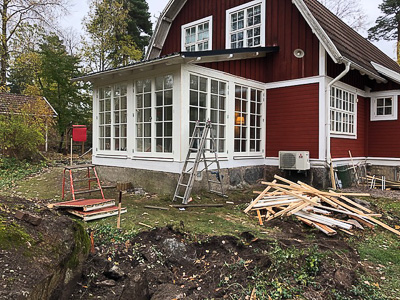 Rött trähus med ny vit glasveranda, där långa spröjsade fönsterpartier och flera höga fönsterdörrar bildar en ljus fasad mot trädgården.