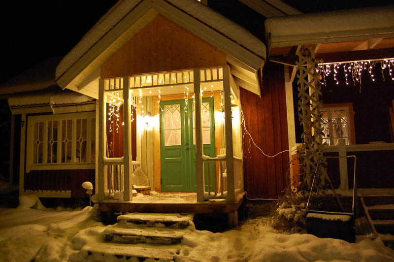 Kvällsbild av farstukvist med grön pardörr, glasfält med kryss/romb-spröjs och julbelysning.