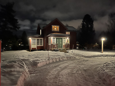 Vinterkväll med rött hus där spröjsade fönster lyser varmt inifrån, och entrépartiet ramas in av glasade sidofönster mot snötäckt gård.