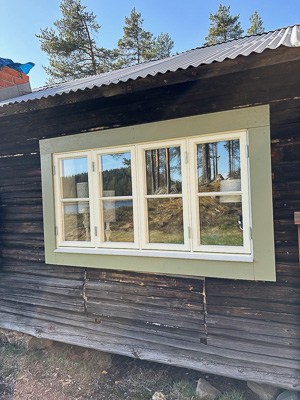 Fyra vita spröjsade fönster i rad på mörk träfasad med grönt foder, där glasen speglar skog och himmel och släpper in mycket ljus.