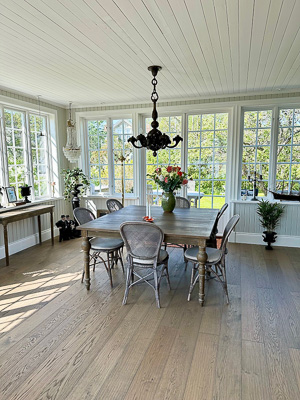 Matplats i glasveranda med spröjsade fönster längs två väggar, där ljuset strilar in och fönstren ramar in utsikten mot trädgården.