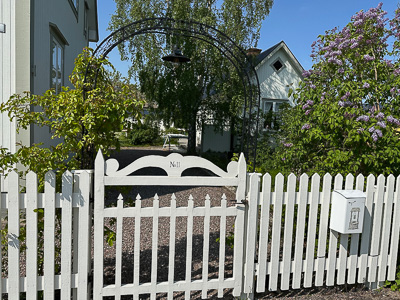 Vit trägrind och staket framför trädgård, med hus i bakgrunden där spröjsade fönster syns mellan grönska och syrener.