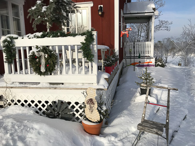 Snötäckt altan med vit glasad dörr och spröjsat fönster intill, där fönsterglasen speglar vinterljuset och ger inblick mot huset.