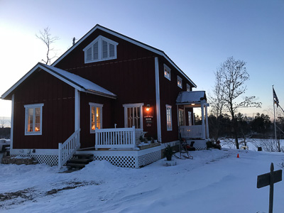 Rött hus i vintermiljö med flera vitmålade spröjsade fönster som lyser varmt inifrån och markerar fasadens fönsterindelning.