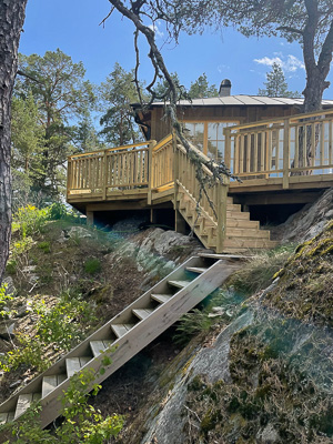 Trähus på klipphäll med stor altan, där fasaden har breda glaspartier och fönster som speglar himmel och tallar och släpper in mycket dagsljus mot terrassen.