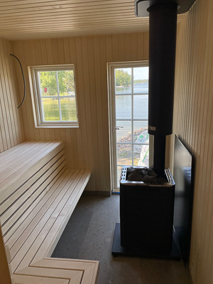 Bastu med ljust träpanel, ett rektangulärt fönster på väggen och en glasad dörr som släpper in ljus och ger utsikt mot vatten och strand utanför.