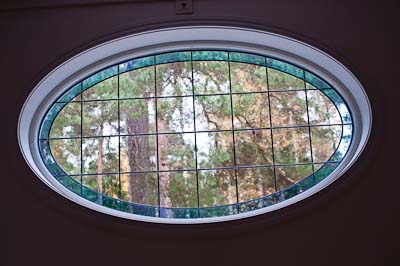 Ovalt blyinfattat fönster sett inifrån, med spröjsade rutor och färgat glas i ytterkanten som ramar in utsikten mot skog utanför.