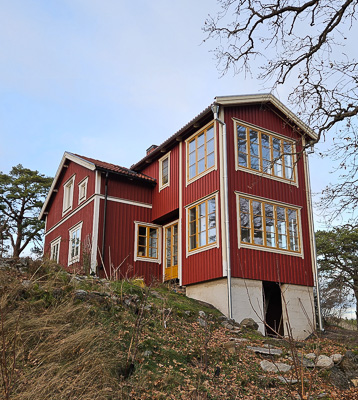 Rött trähus med gulmålade spröjsade fönster, där hörnbyggnaden domineras av två stora fönster med 10 glasrutor vardera (5×2) samt ett sidofönster med 6 glasrutor (2×3), med himmel och träd som speglas i rutorna.
