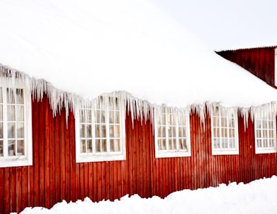 Röd träbyggnad vintertid med vita spröjsade fönster, där långa istappar hänger från ett snötäckt tak. Snön ligger djup längs fasaden och bilden förmedlar kyla, vinterstämning och traditionell svensk industrimiljö.