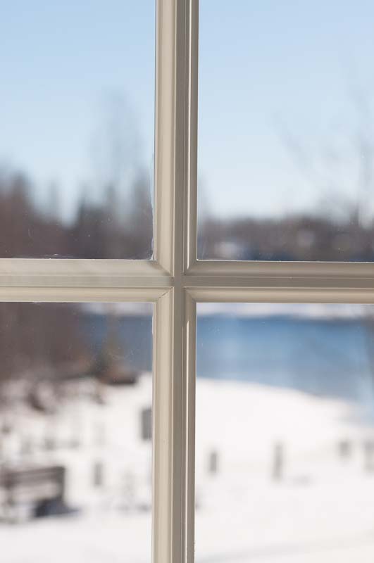 Närbild av ett vitmålat spröjsat fönster med enkelglas, där vinterlandskap och vatten syns suddigt genom rutorna.