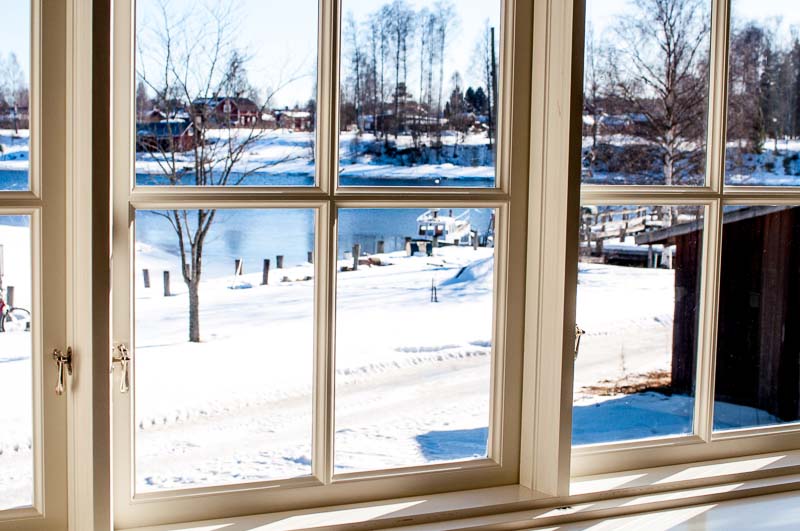 Vitmålat fönsterparti med spröjsade enkelglasfönster och lösa innerbågar, där snötäckt mark, vatten och bebyggelse syns utanför i dagsljus.