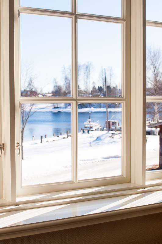 Vitmålat spröjsat fönster med enkelglas och lösa innerbågar, där vinterljus faller in över fönsterbänken och snötäckt landskap syns utanför.