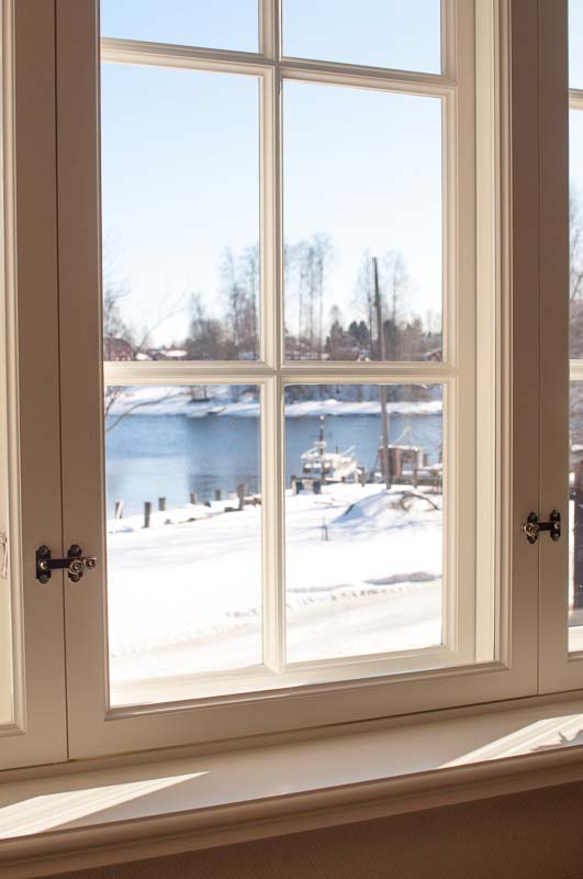 Vitmålat spröjsat fönster med enkelglas och lösa innerbågar, där ett snötäckt landskap och vatten syns genom rutorna i dagsljus.