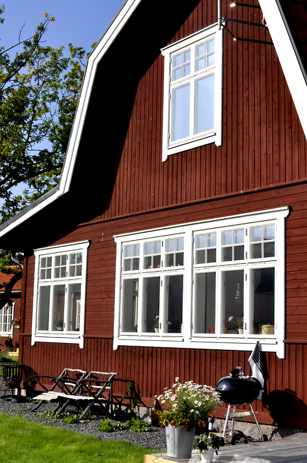 Rött trähus med vitmålade spröjsade sexluftsfönster och gavelfönster, beläget i grön trädgård med utemöbler och grill.