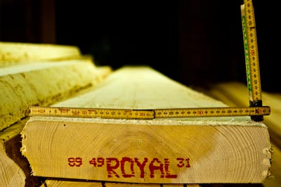 Närbild av en grov träplanka med ändträ i fokus, där måttstockar ligger utlagda i vinkel för att mäta dimensionerna. På träets ändyta syns en röd stämpel med texten “ROYAL”. Bilden förmedlar kvalitetskontroll och noggrann mätning av råvirke i en snickerimiljö.