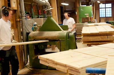 Tre snickare arbetar i en träverkstad med en stor grön industrimaskin. En av dem matar in långa träbrädor i maskinen medan andra övervakar arbetet. I förgrunden syns högar av färdigbearbetade träskivor staplade på bockar. Alla bär arbetskläder och hörselskydd, och miljön präglas av högt tempo och pågående industriell träbearbetning i fabriken.