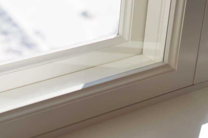 Närbild av ett vitmålat fönster med enkelglas och lös innerbåge, som visar fönstrets karm, smyg och fönsterbänk i dagsljus.