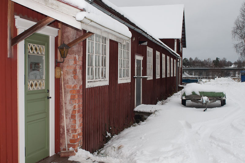 Röd träbyggnad med vita fönster och snötäckt tak under vintertid. I förgrunden syns en grön ytterdörr med spröjsade glasrutor och en tegelmur vid entrén. Marken är täckt av djup snö, och längs byggnaden leder en snöig gång mot en liten båt vid vattnet i bakgrunden. Stämningen är stillsam och vintrig.