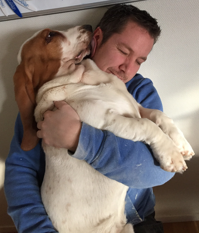 Person som sitter och kramar en stor hund i famnen, där hunden slickar personen i ansiktet i en varm och lekfull stund.
