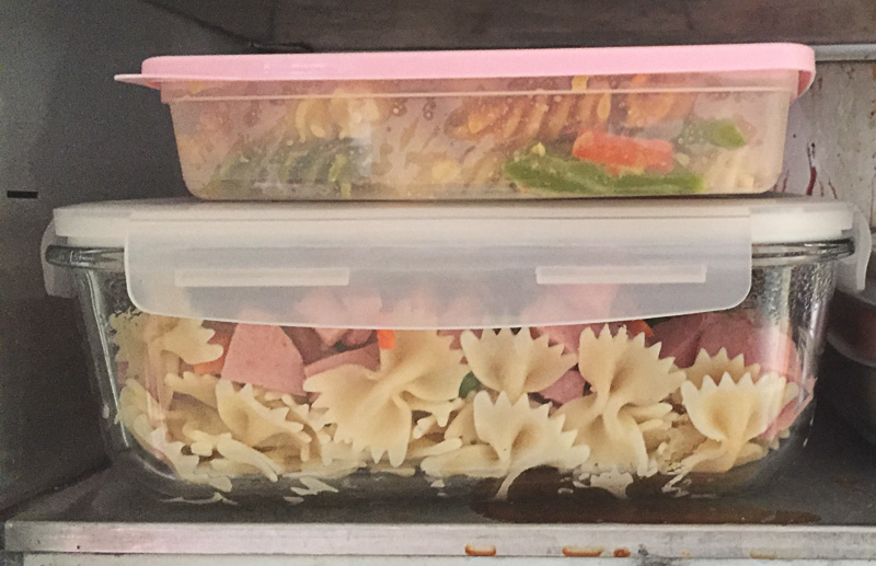 Två matlådor staplade i ett kylskåp, fyllda med pastasallad med farfalle, grönsaker och skinka.