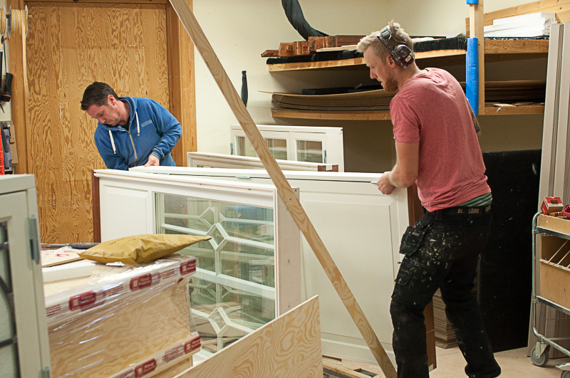 Två snickare i verkstadsmiljö som arbetar med ett stort vitmålat fönsterparti med spröjs, omgivet av träskivor, verktyg och material under pågående produktion.