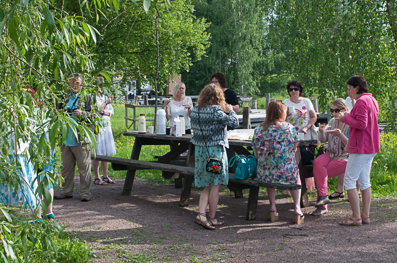 Grupp besökare samlade runt ett picknickbord utomhus i grönskande miljö, där de fikar och samtalar under ett studiebesök.