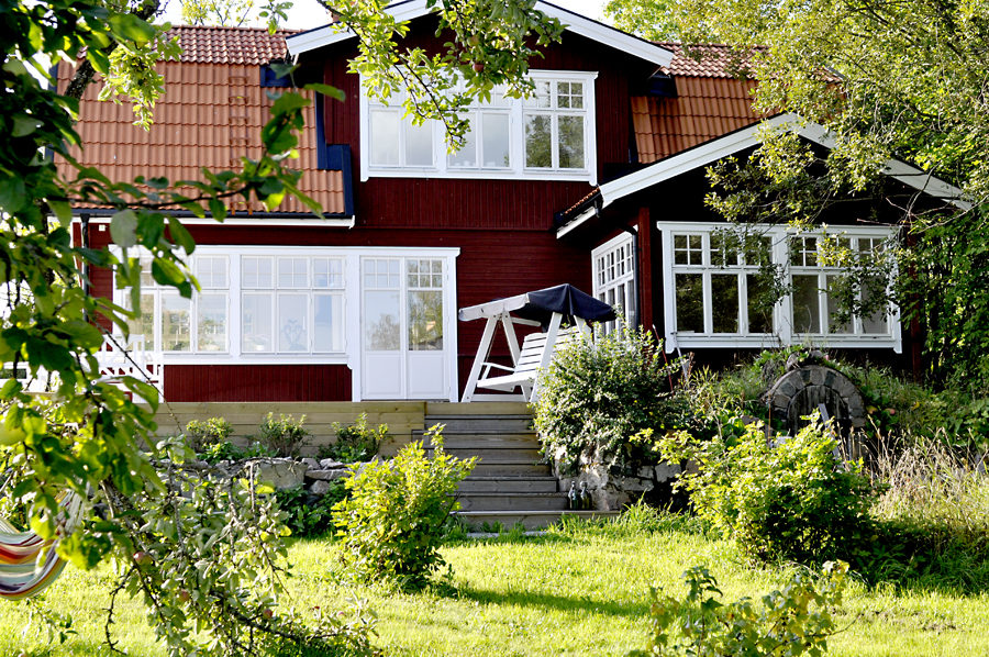 Rött trähus med vitmålade spröjsade fönster och veranda, omgivet av grönskande trädgård och träd, sett från en lägre vinkel i dagsljus.