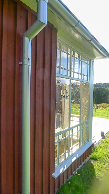 Sidoparti på glasveranda med spröjsat rutnät och lång fönsterbänk, röd stående panel utvändigt.