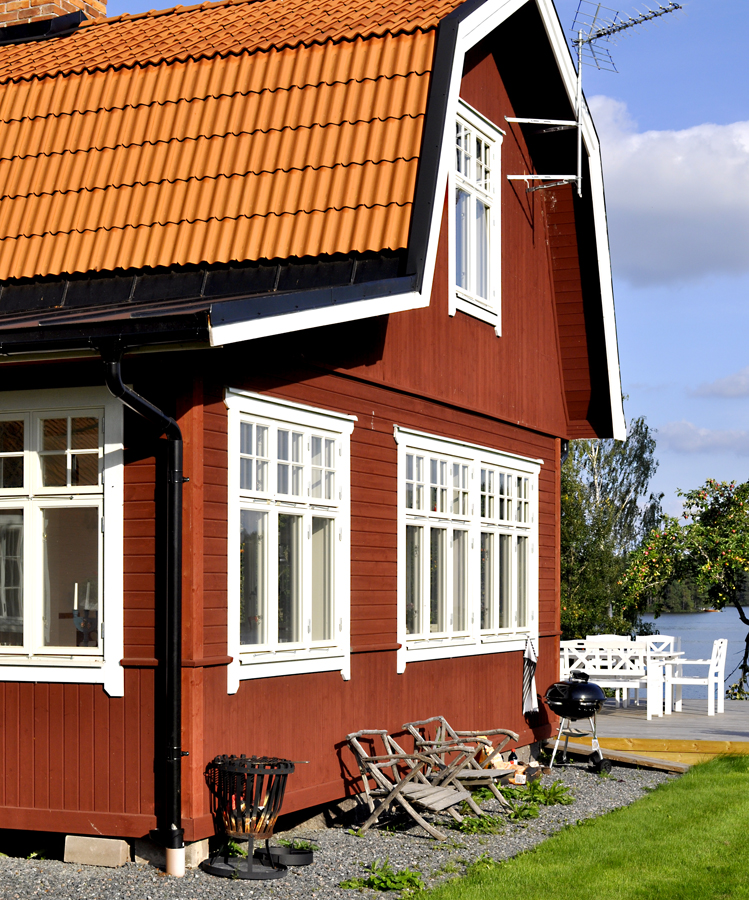 Rött trähus med vitmålade spröjsade fönster och gavelfönster, täckt av tegeltak, beläget i grön trädgård med altan och utemöbler.