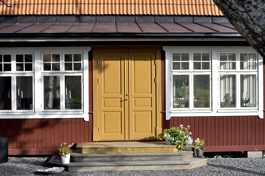 Entré med gul pardörr och speglar, flankerad av vitmålade spröjsade fönster, monterad i en röd träfasad med liten trappa och blomkrukor.