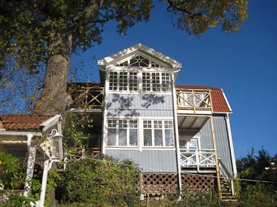 Blå fasad med verandatillbyggnad, spröjsade fönster i rutnät och dekorativt överljus.