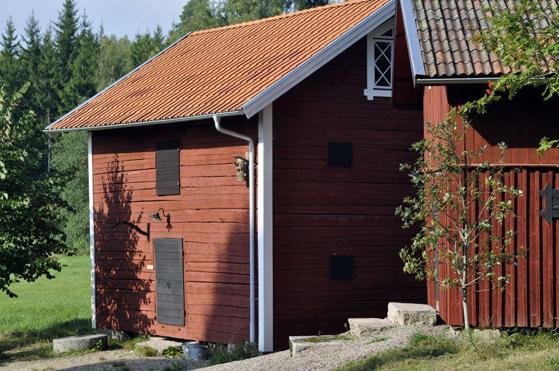 Rödmålad träbyggnad med tegeltak och enkla fönsterluckor, belägen i lantlig miljö med grönska och träd runt omkring.