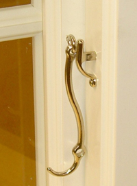 Långt svängt fönsterhandtag i mässing på vit karm intill glasruta.