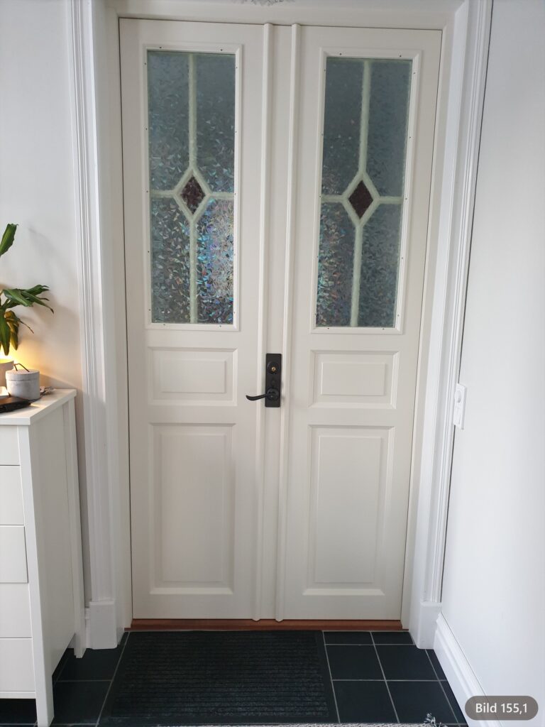Röd pardörr med 2 höga glasrutor i strukturerat glas och diamantspröjs med röd glasromb som släpper in ljus i hallen.
